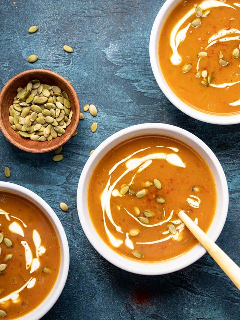 Three bowls of easy pumpkin soup garnished with sour cream and pumpkin seeds, on a blue background.