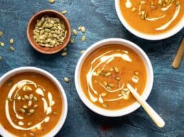 Three bowls of Easy Pumpkin Soup garnished with sour cream and pepitas, a small bowl of pepitas on the side.