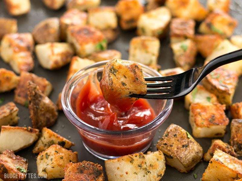 One parmesan roasted potato being dipped into a small dish of ketchup, surrounded by more roasted potatoes