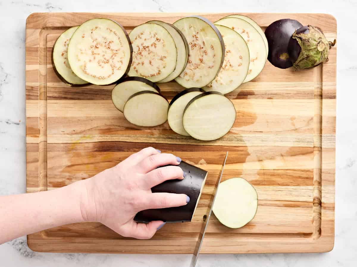 A hand slicing an eggplant into rounds on a wooden cutting board.