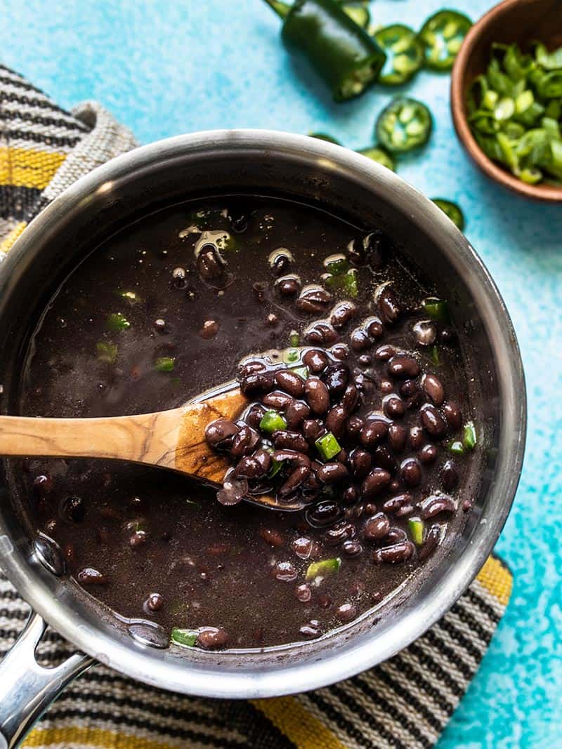 A wooden spoon lifting some quick seasoned black beans out of a sauce pot, jalapeños and green onion on the side