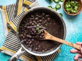 A pot of quick seasoned black beans, with a wooden spoon, jalapeños and green onion on the side