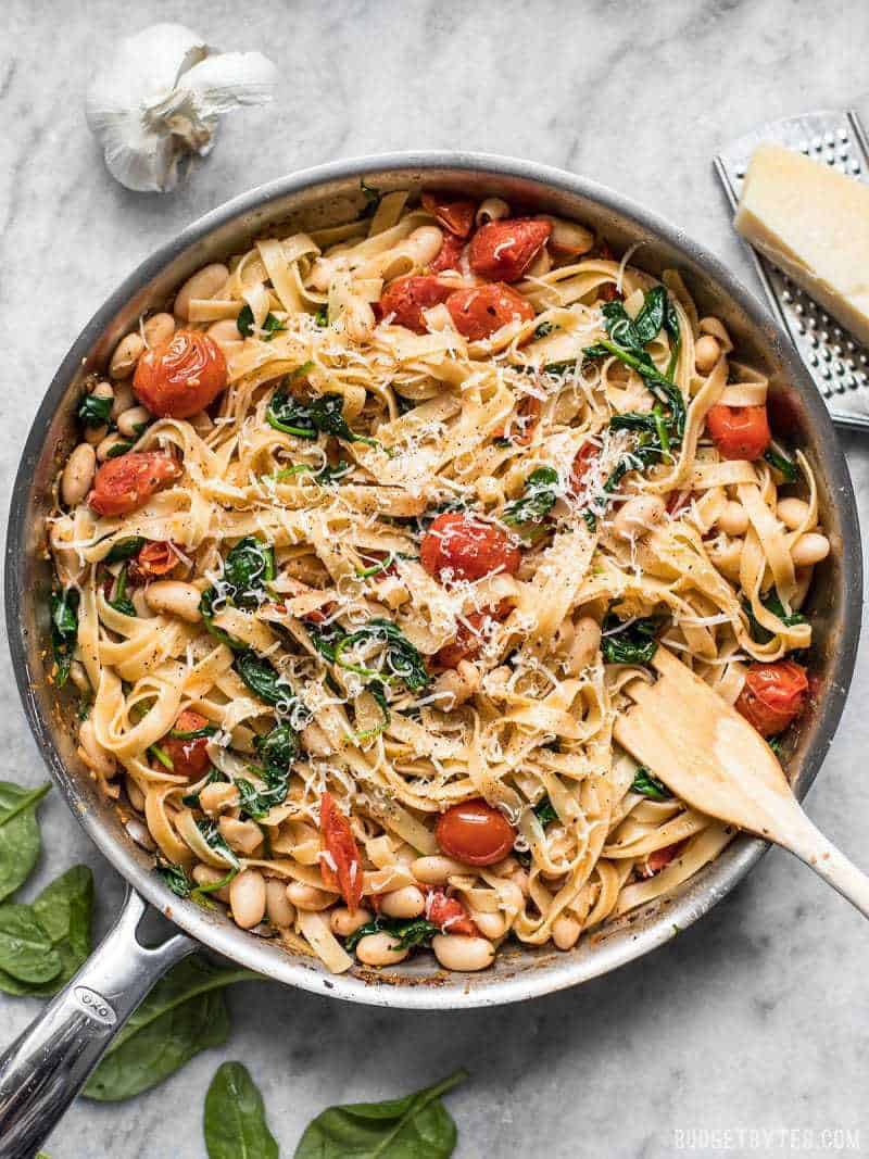 Overhead view of the skillet full of tuscan white bean pasta with a wooden pasta fork in the side, garlic and Parmesan near the skillet