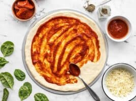 Pizza sauce being spread onto a stretched pizza dough with toppings on the sides