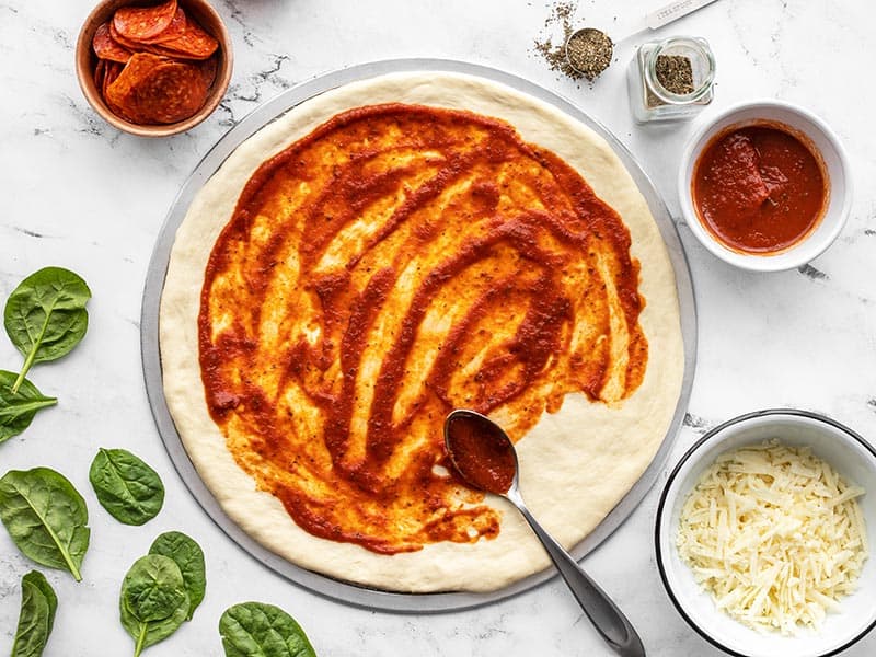 Pizza sauce being spread onto a stretched pizza dough with toppings on the sides