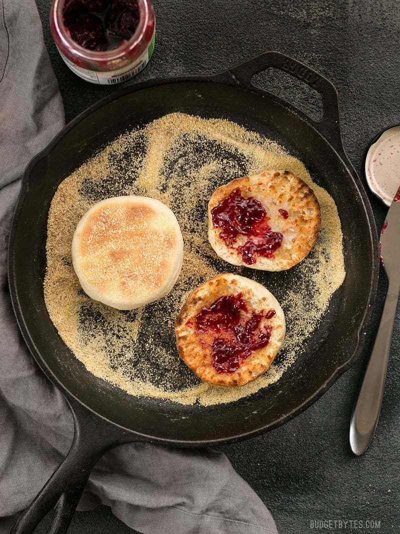 Homemade English muffins in a cast iron skillet full of cornmeal, jam smeared on a couple toasted muffin halves.