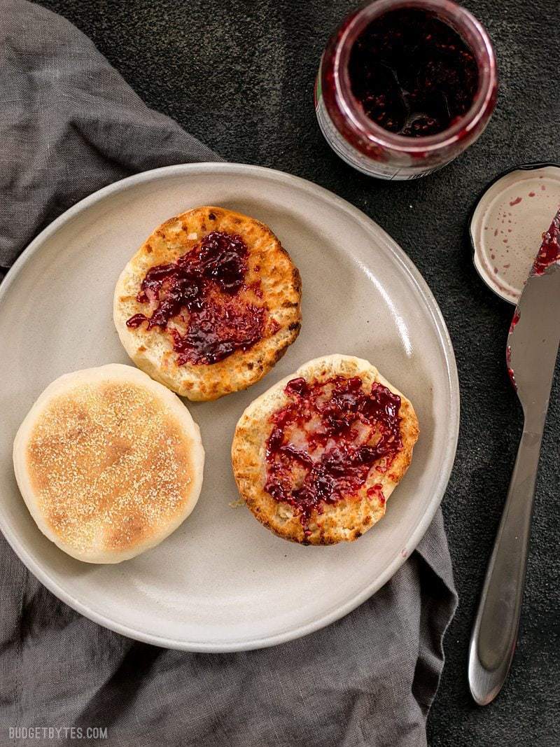 Two English muffins on a plate, one sliced open and toasted, smeared with raspberry jam.