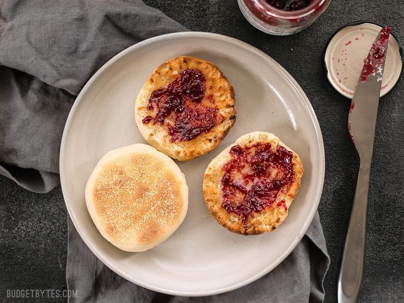 Homemade English muffins are fun to make, delicious, and cost just pennies each. Make this your next weekend project! Step by step photos. BudgetBytes.com Sliced and toasted english muffins on a plate, one smeared with raspberry jam.