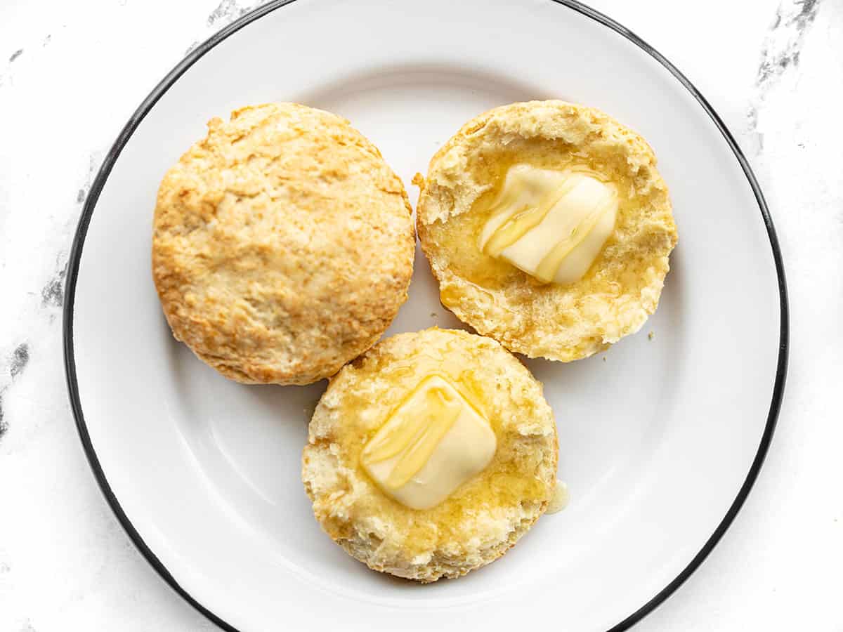 Two butter biscuits on a plate, one sliced open and topped with butter and honey