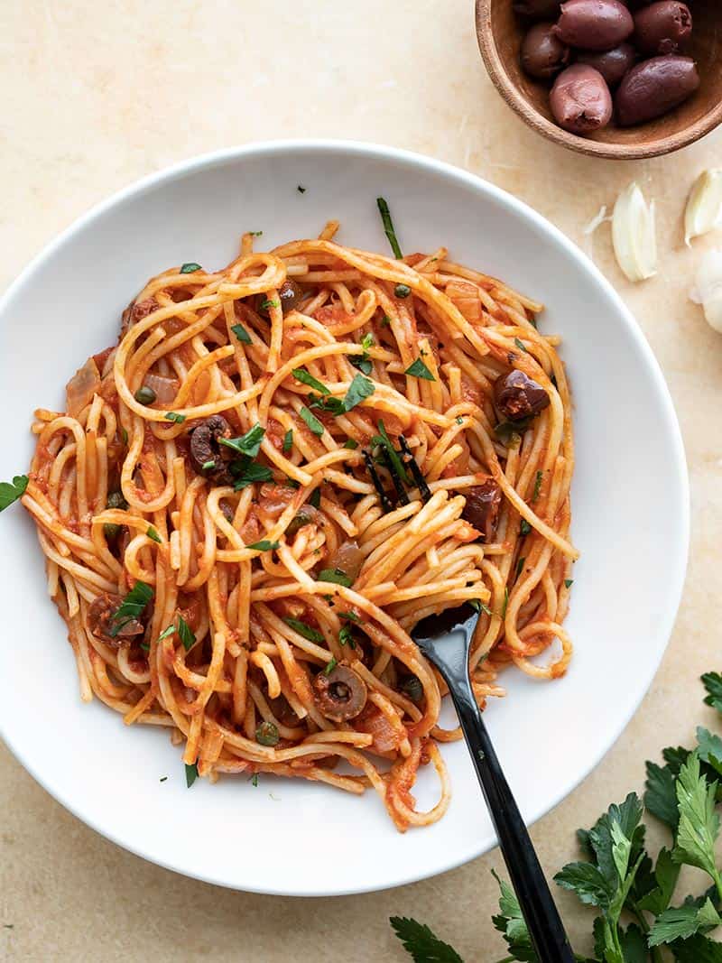 A large bowl of pasta puttanesca with a fork twirled in the center and a bowl of olives on the side.