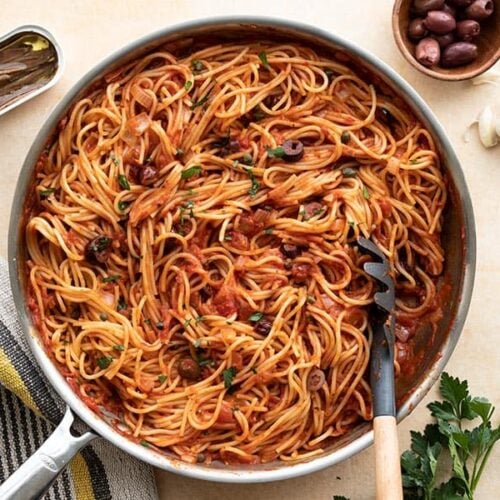 A skillet full of pasta puttanesca with anchovies and olives on the side