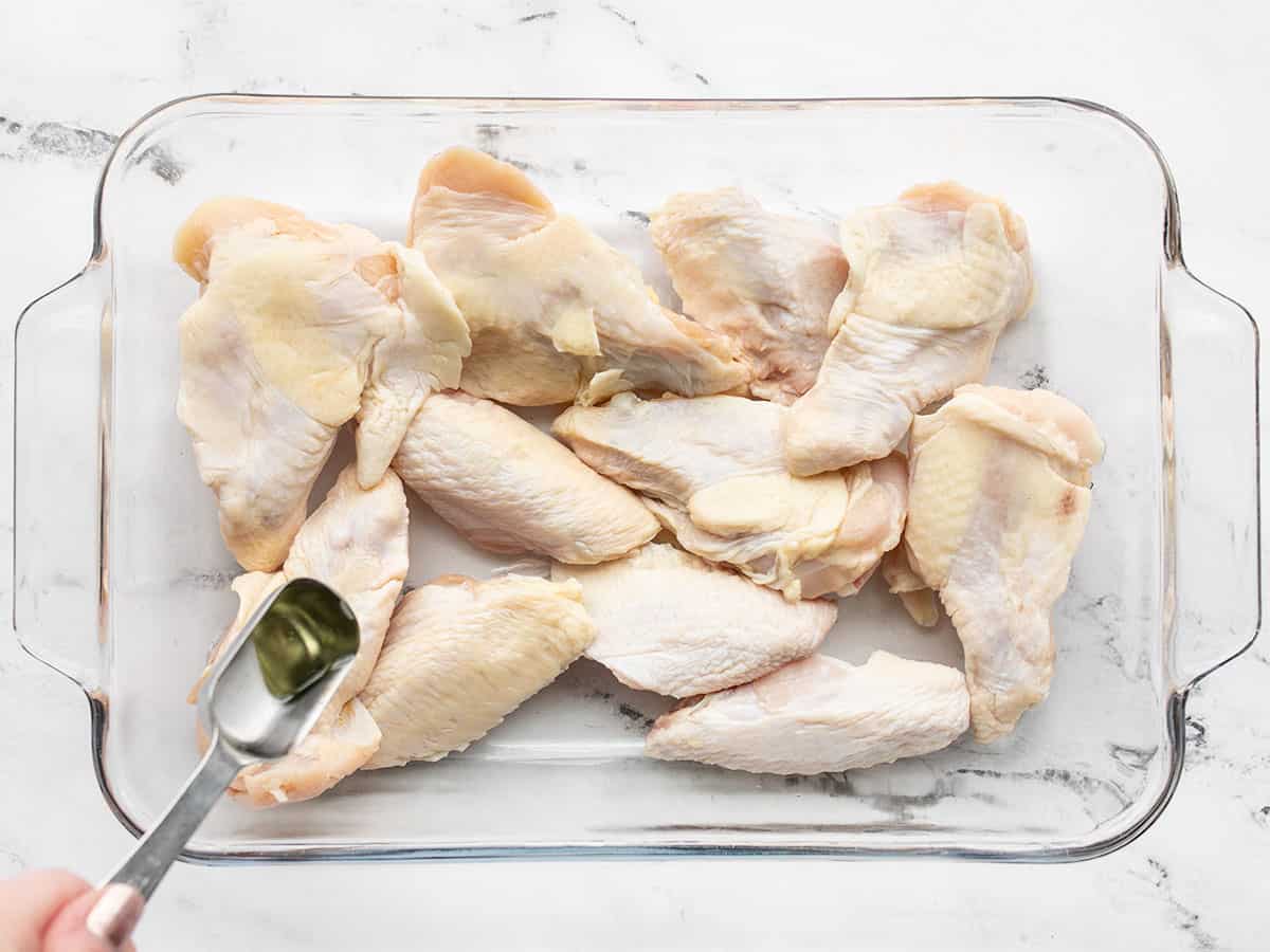 Raw chicken wings in a casserole dish being drizzled with cooking oil