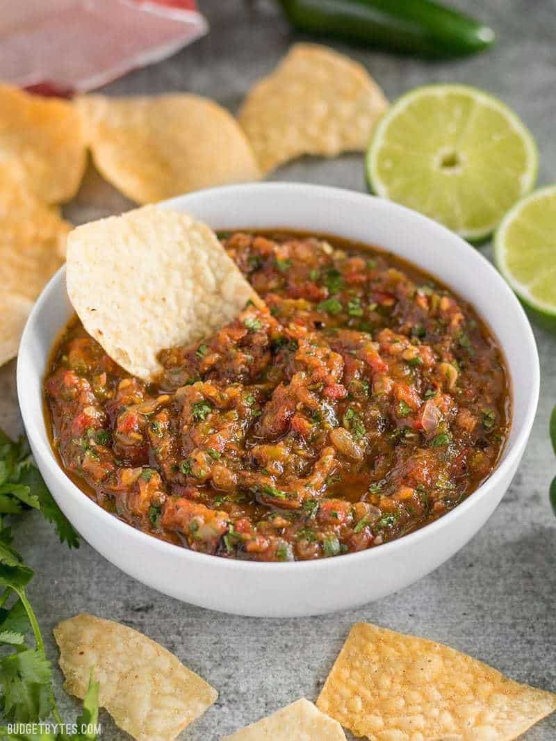 Fire Roasted Salsa in white bowl with tortilla chip dipped in. Limes on side. 