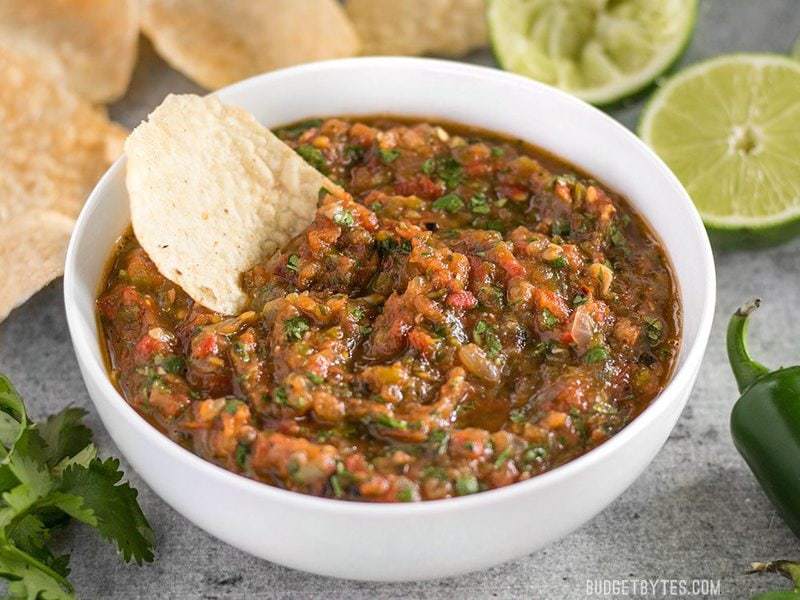 Fire Roasted Salsa in white bowl with tortilla chip dipped in. Limes and Jalapeño on the side. 