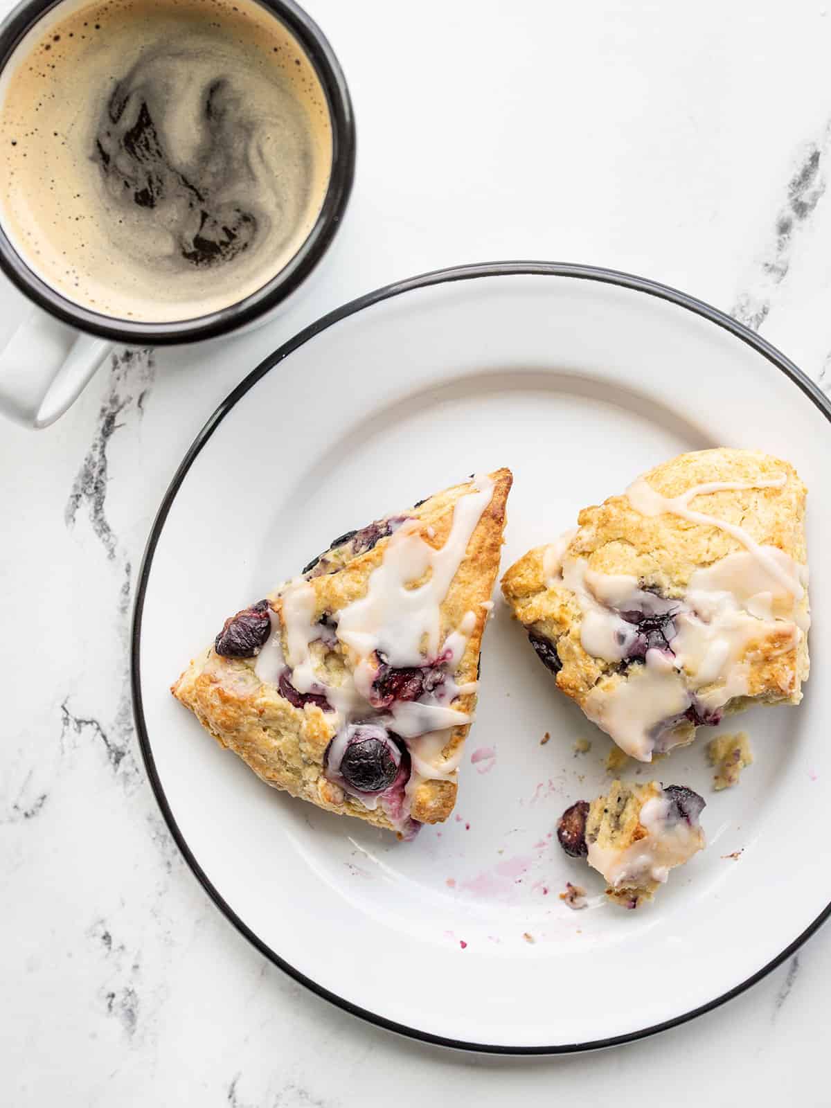 Two lemon blueberry scones on a plate with a mug of coffee on the side