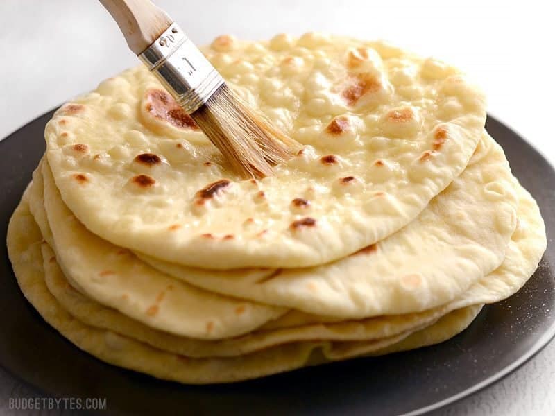 A stack of freshly baked naan on a plate being brushed with butter. 