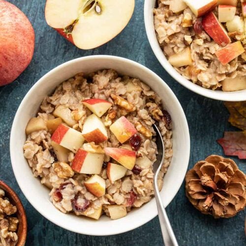 Two bowls of autumn fruit and nut oatmeal surrounded by apples, leaves, and pinecones