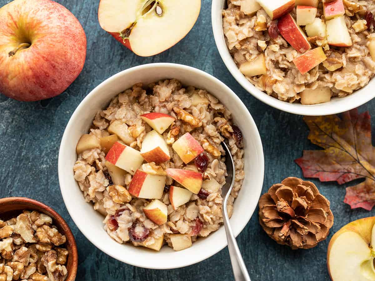 Two bowls of autumn fruit and nut oatmeal surrounded by apples, leaves, and pinecones 