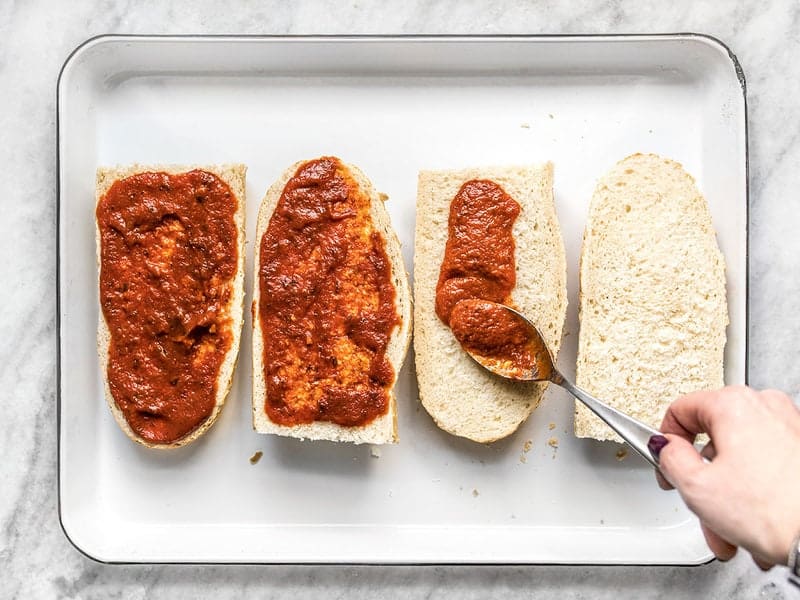 A hand smearing pizza sauce on pieces of French Bread with a spoon