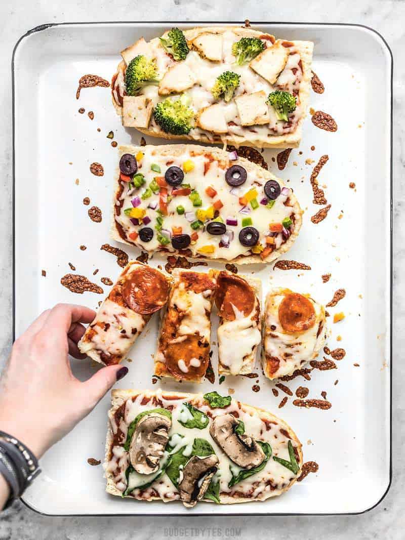 French Bread Pizza is the perfect budget-friendly fast and easy weeknight dinner. Customize the toppings to fit your taste buds or what you have on hand! Budgetbytes.com Four French Bread Pizzas on a baking sheet with a hand taking one piece
