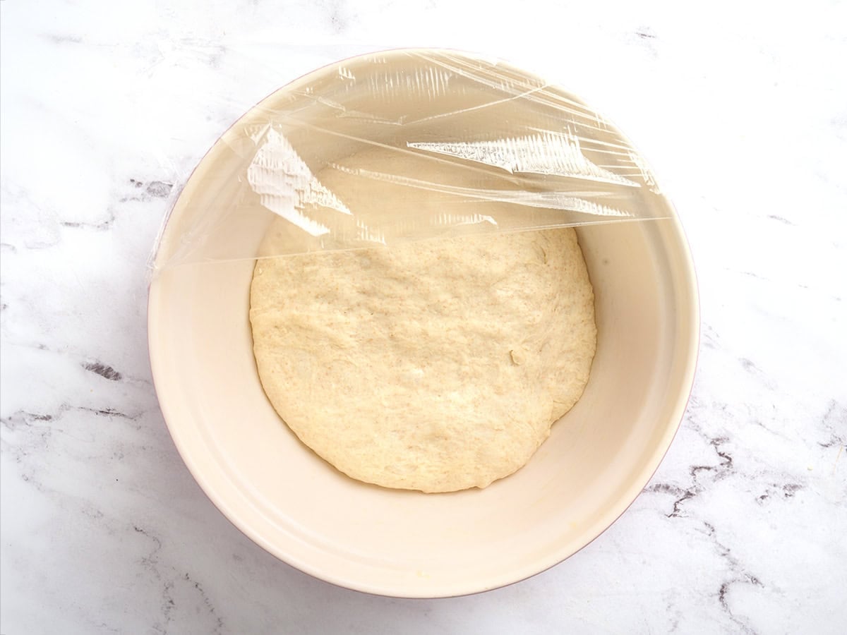 Pita bread dough in a bowl after proofing and covered with plastic wrap.