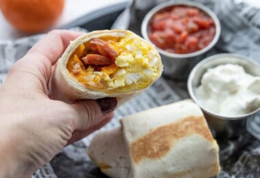 A hand holding half of a breakfast burrito with the open cut side facing the camera.