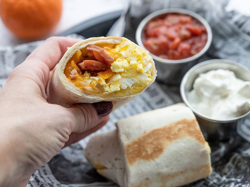 A hand holding half of a breakfast burrito with the open cut side facing the camera.