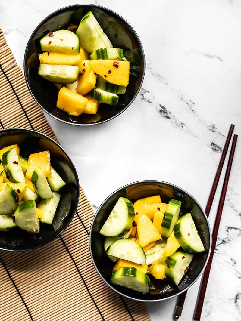 Three black ceramic bowls with Cucumber mango salad on a marble table with a bamboo mat and chopsticks.