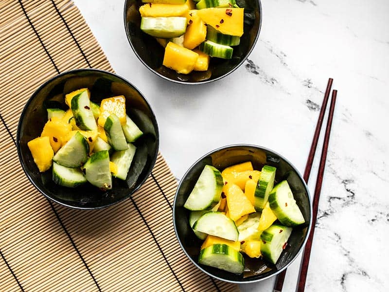 Three bowls of cucumber mango salad on a bamboo mat with chopsticks.