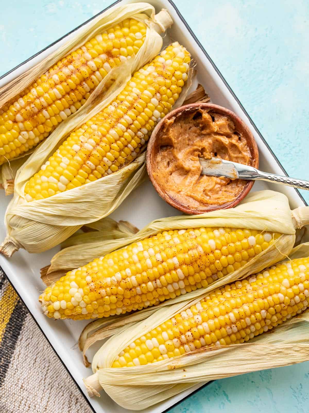 Oven roasted corn half shucked and smeared with honey chili butter on a white tray with a bowl of butter