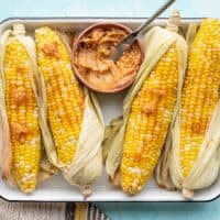 Oven roasted corn on a tray, partially shucked, with a bowl of honey chili butter