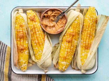 Oven roasted corn on a tray, partially shucked, with a bowl of honey chili butter
