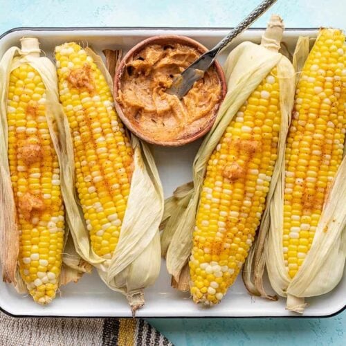 Oven roasted corn on a tray, partially shucked, with a bowl of honey chili butter