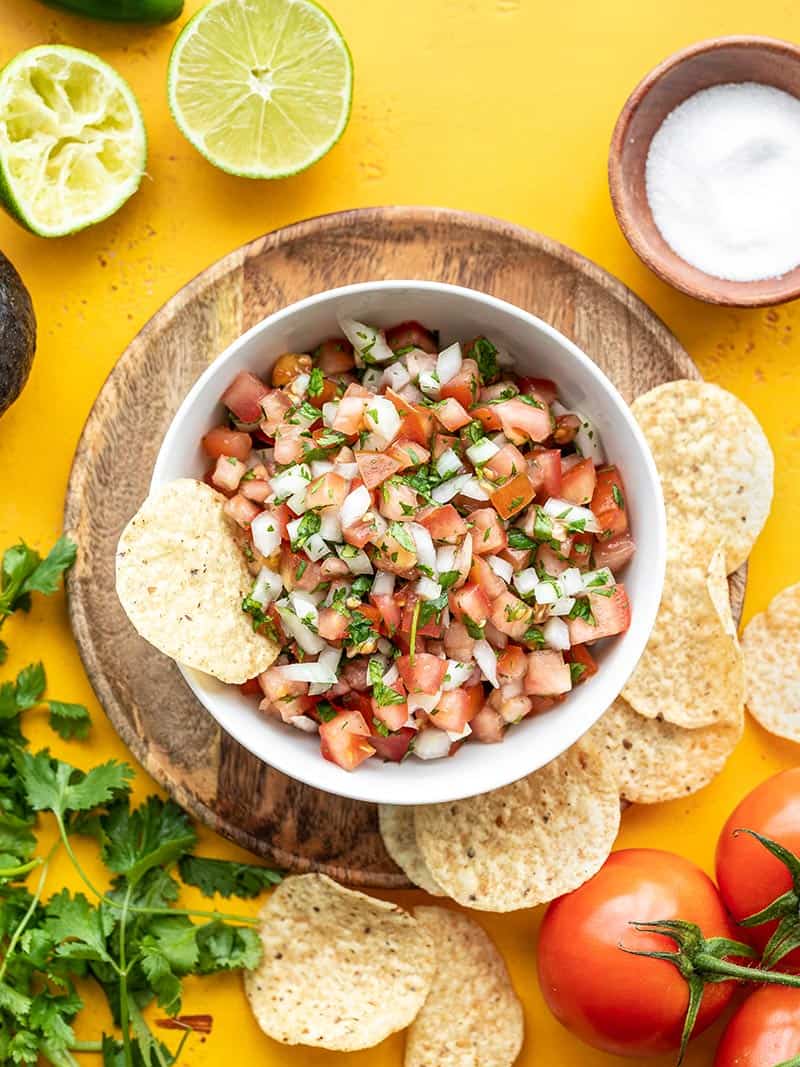 A bowl of pico de gallo surrounded by chips, tomatoes, limes, cilantro, and salt.