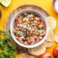 Overhead view of a bowl of pico de gallo surrounded by tortilla chips, salt, tomatoes, cilantro, and limes