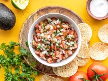 Overhead view of a bowl of pico de gallo surrounded by tortilla chips, salt, tomatoes, cilantro, and limes