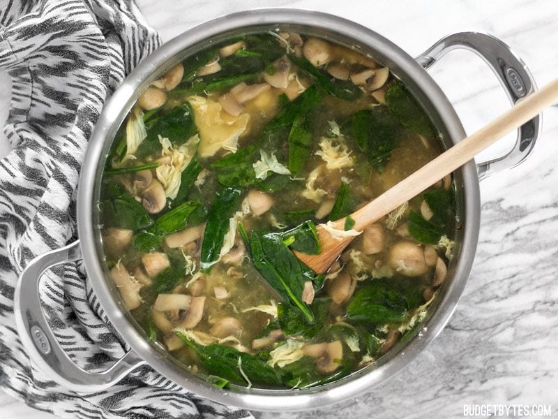 Finished pot of homemade easy egg drop soup with spinach, mushrooms, and a wooden spoon in the side