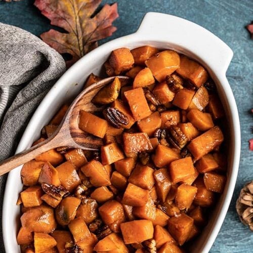 Maple Roasted Sweet Potatoes in a casserole dish with a wooden spoon on a blue background with leaves and pine cones on the side.