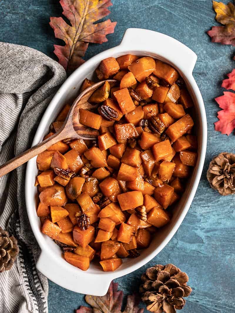 Overhead view of a casserole dish full of Maple Roasted Sweet Potatoes with pecans with a wooden spoon on a deep blue background