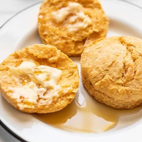 Close up of two sweet potato biscuits on a plate, one open, smeared with butter, and drizzled with maple syrup