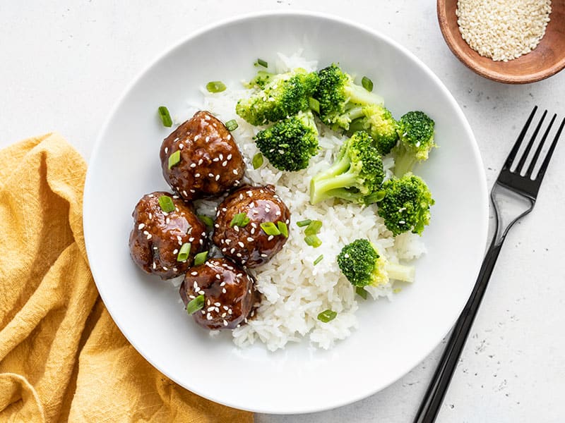 Finished teriyaki meatball bowls with a fork, yellow napkin, and bowl of sesame seeds.