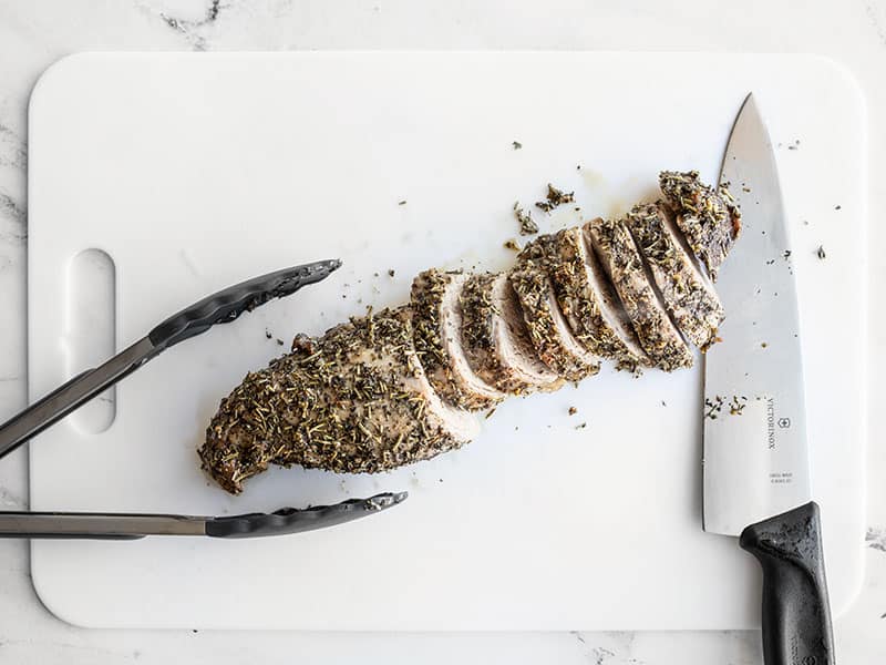 Herb Roasted Pork Tenderloin being sliced on a white cutting board.