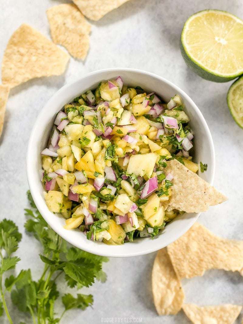 A bowl of pineapple salsa viewed from above with tortilla chips, cilantro, and limes on the side. One chip stuck in the bowl.