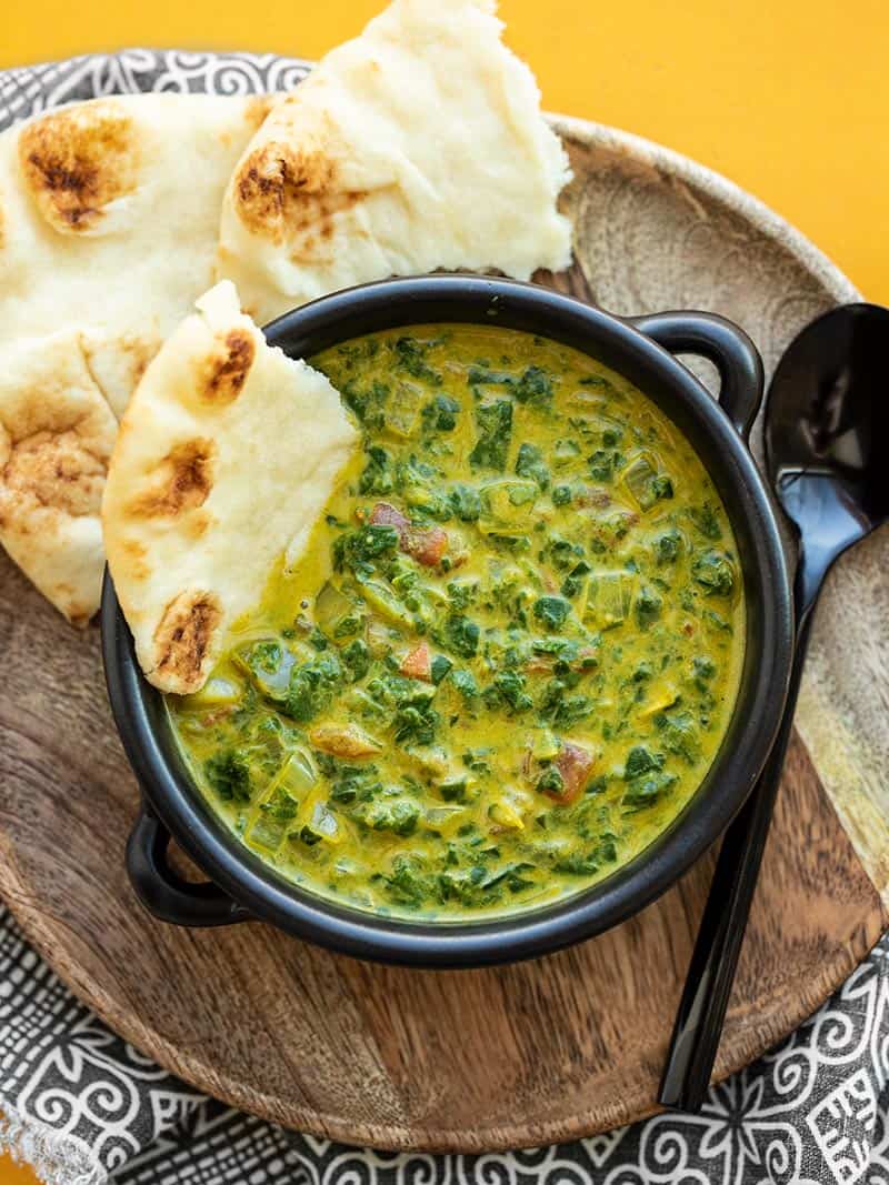 A black bowl full of Indian Creamed Spinach on a wooden plate with a piece of naan dunked into the bowl
