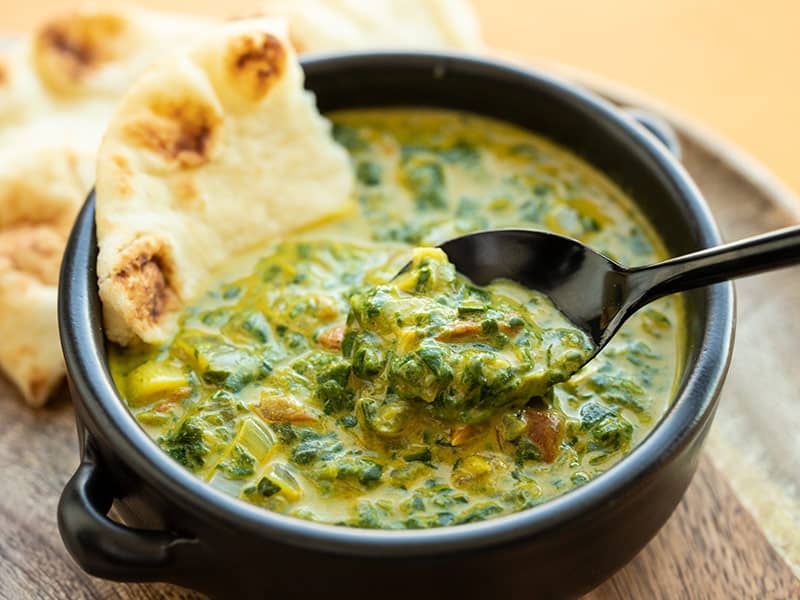 Front view of a bowl full of Indian style creamed spinach with a spoon lifting a bite and a piece of naan stuck in the side of the bowl