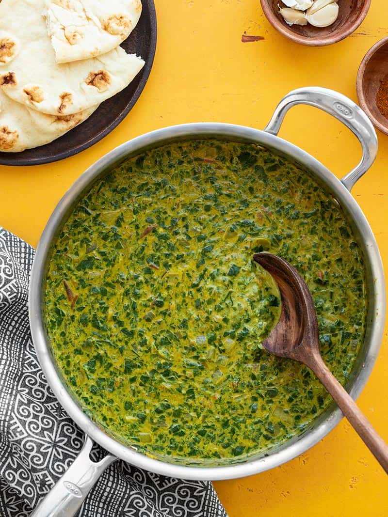 A finished skillet full of Indian Style Creamed Spinach with naan on the side and a wooden spoon in the center of the skillet