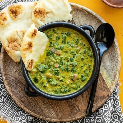 A black bowl with Indian Creamed Spinach on a wooden plate with a piece of naan in the side of the bowl