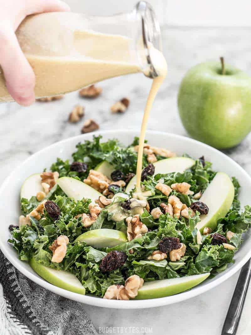 Dijon vinaigrette being poured from a glass bottle onto a bowl of Apple Dijon Kale Salad 