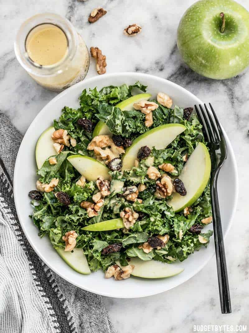 A bowl of Apple Dijon Kale Salad with a jar of dressing on the side and a whole green Granny smith apple