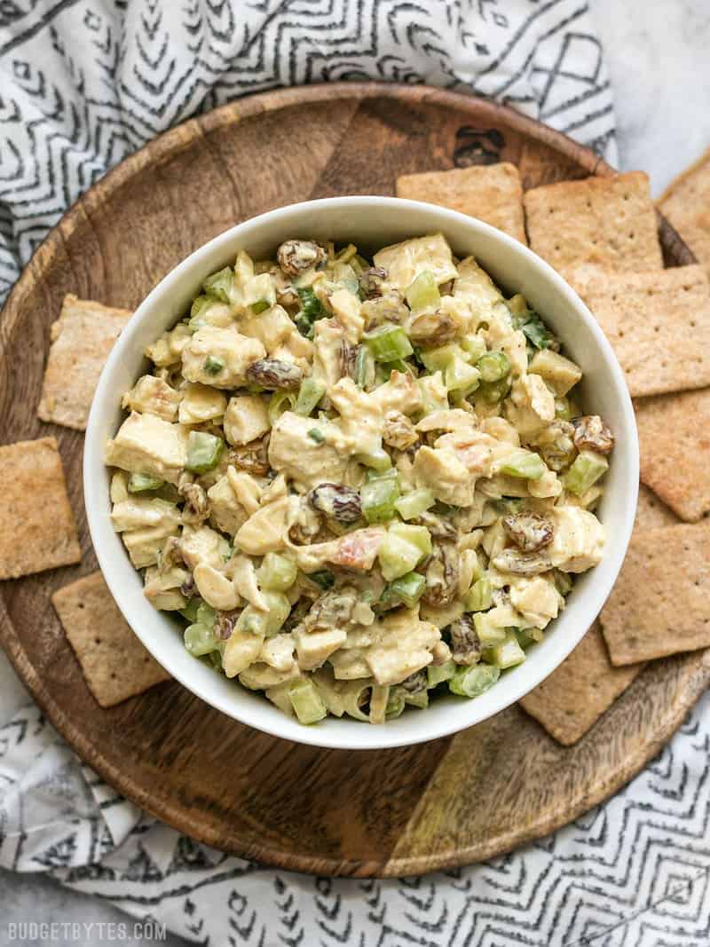 A bowl of Curry Chicken Salad sitting on a wooden plate, surrounded by crackers. 